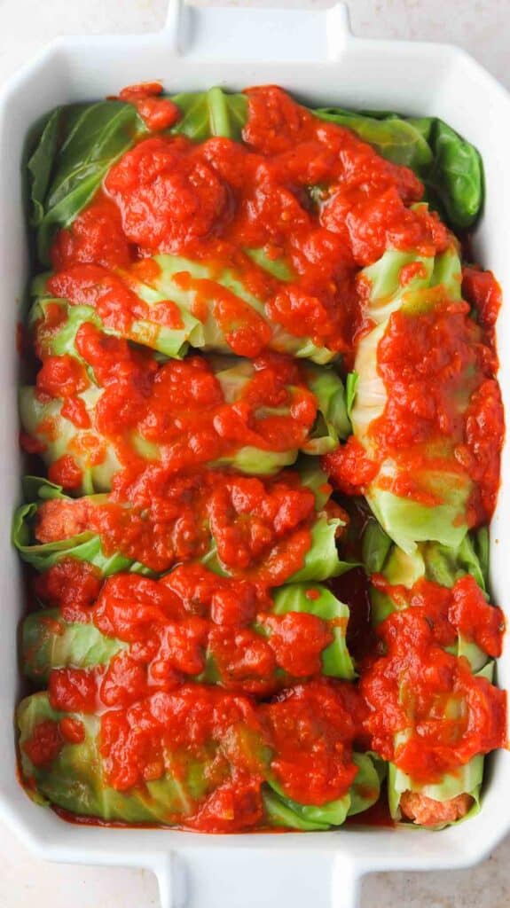 Raw stuffed cabbage in a white casserole dish with red sauce on top.