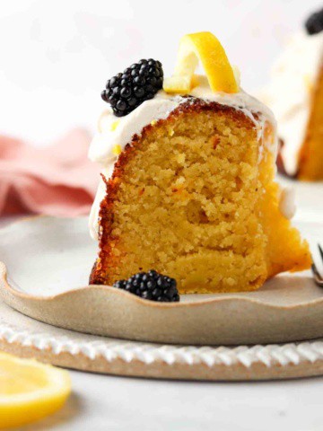 Close up photo of a piece of cake on a plate with blackberries and lemons on top.