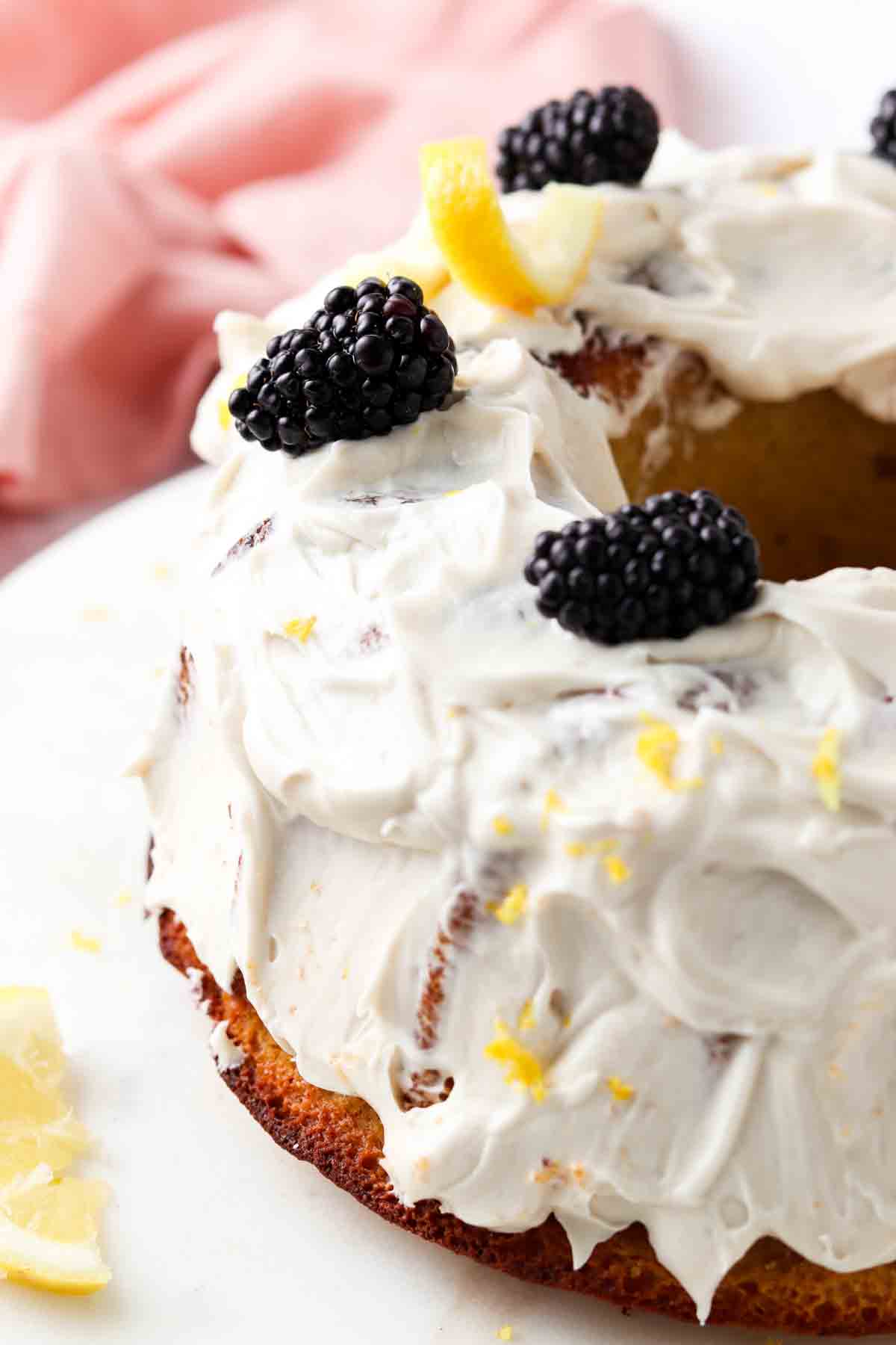 Pound cake with white frosting and blackberries and lemon rinds on top with a pink napkin next to it. 