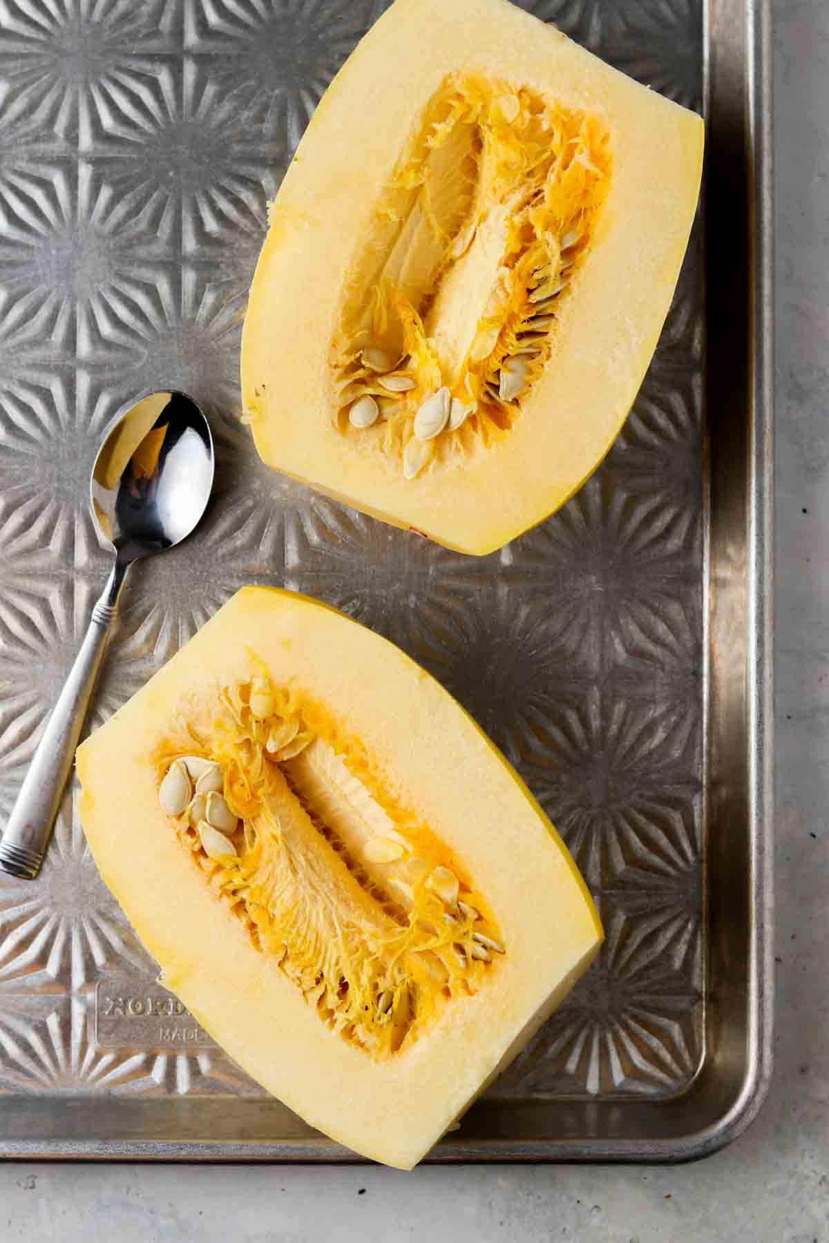 Uncooked spaghetti squash sliced in half on a baking sheet with a spoon next to it.