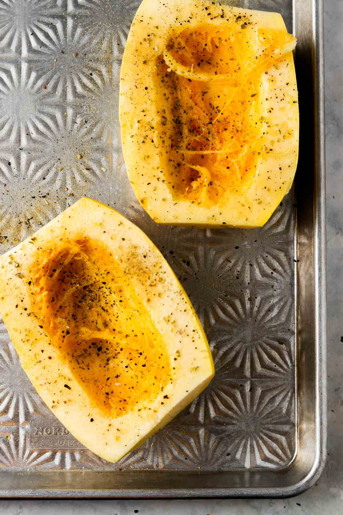 Uncooked spaghetti squash sliced in a half on a silver baking sheet seasoned.