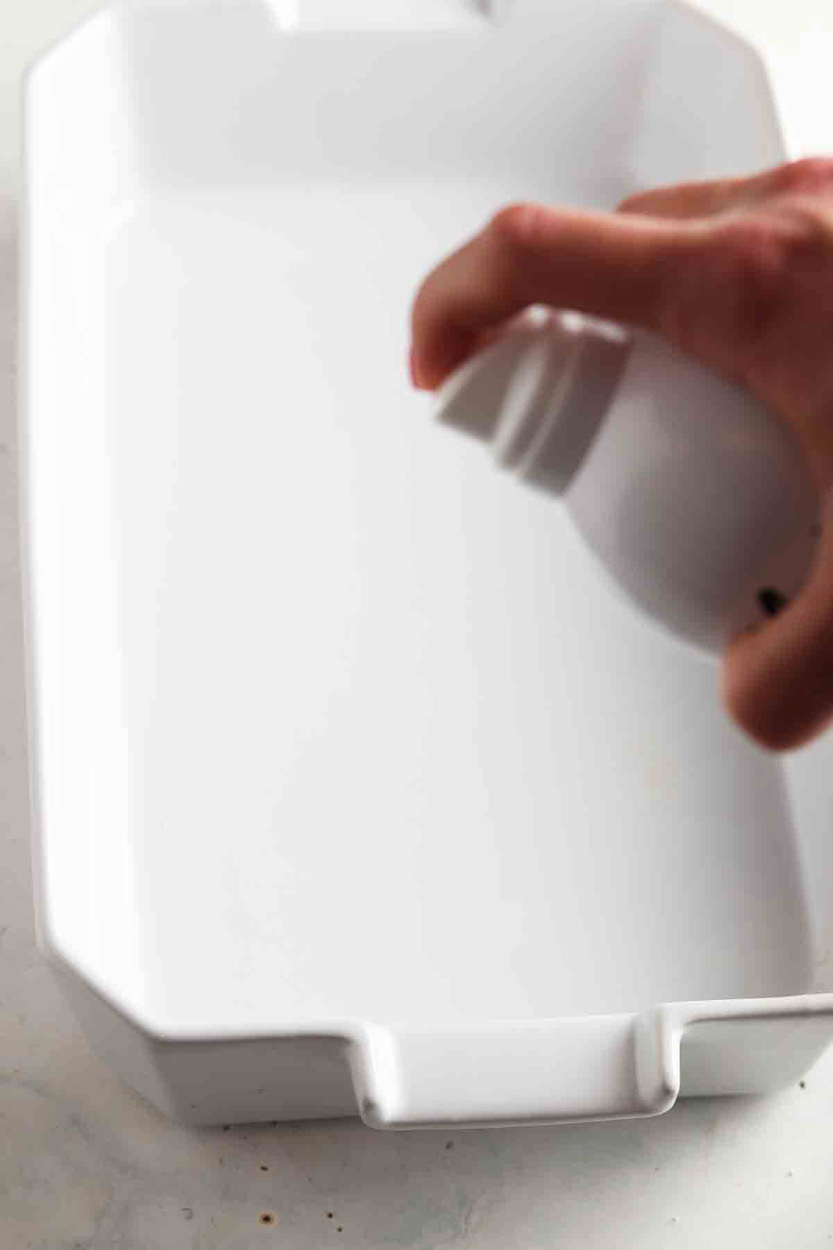 Empty white baking dish being sprayed with cooking oil.