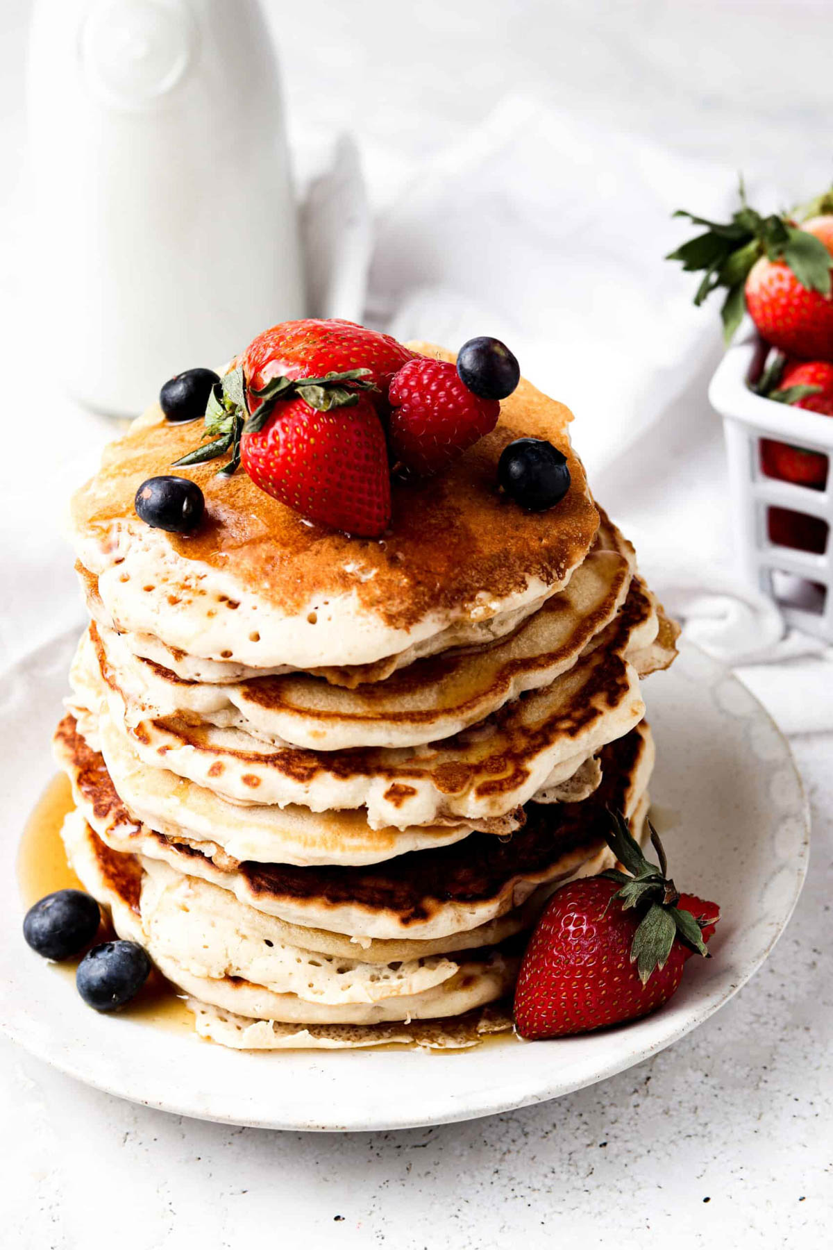 A stack of AIP pancakes on a white plate and topped with syrup and fresh strawberries and blueberries. 