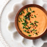 Orange breakfast burrito sauce in a small wooden bowl with sitting in a flower white bowl with fresh chives and red pepper flakes on top.