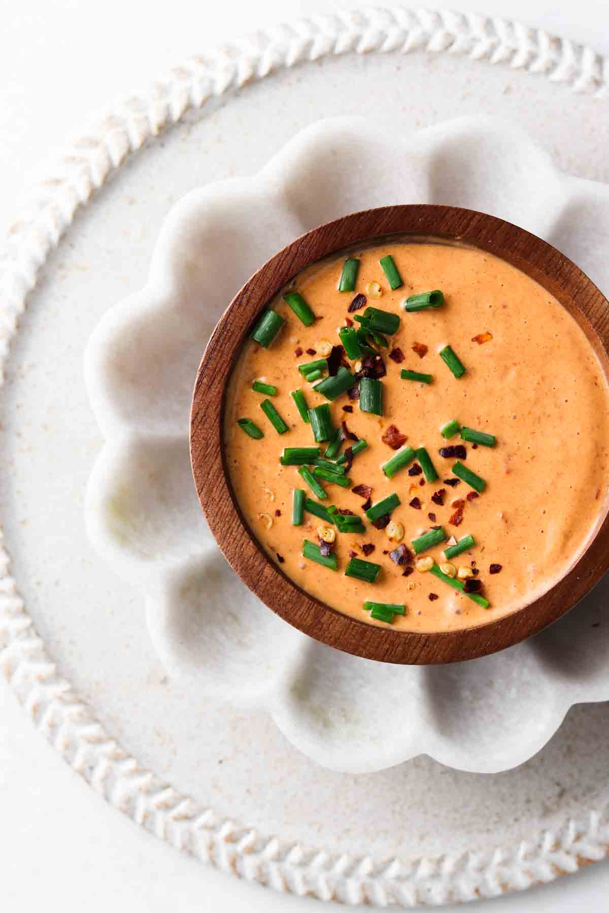 Orange breakfast burrito sauce in a small wooden bowl with sitting in a flower white bowl with fresh chives and red pepper flakes on top.