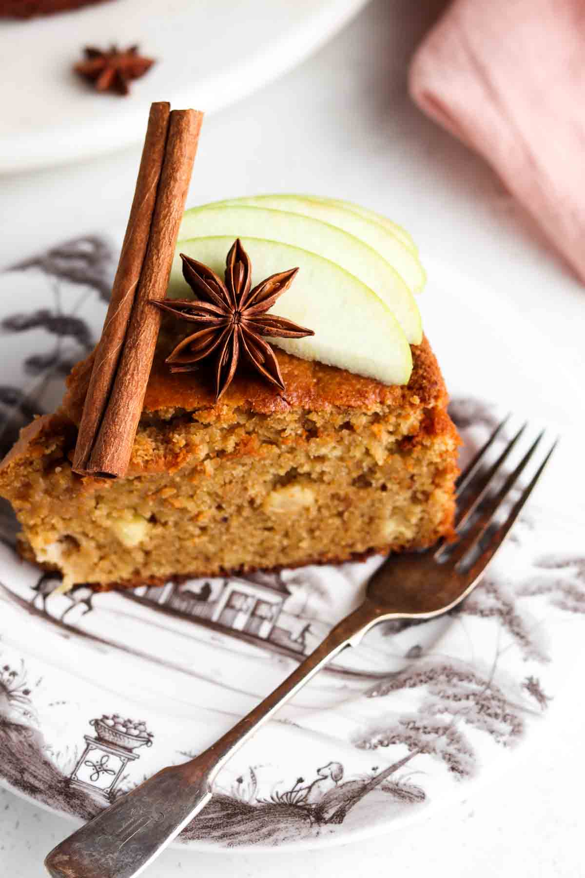 A slice of Gluten Free Apple Cake on a plate by itself with a fork on the side and green apples on top.