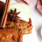 A slice of Gluten Free Apple Cake on a white platter with green apples, a cinnamon stick and anise star on top.