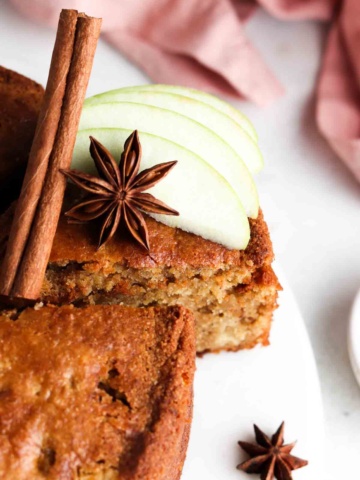 A slice of Gluten Free Apple Cake on a white platter with green apples, a cinnamon stick and anise star on top.