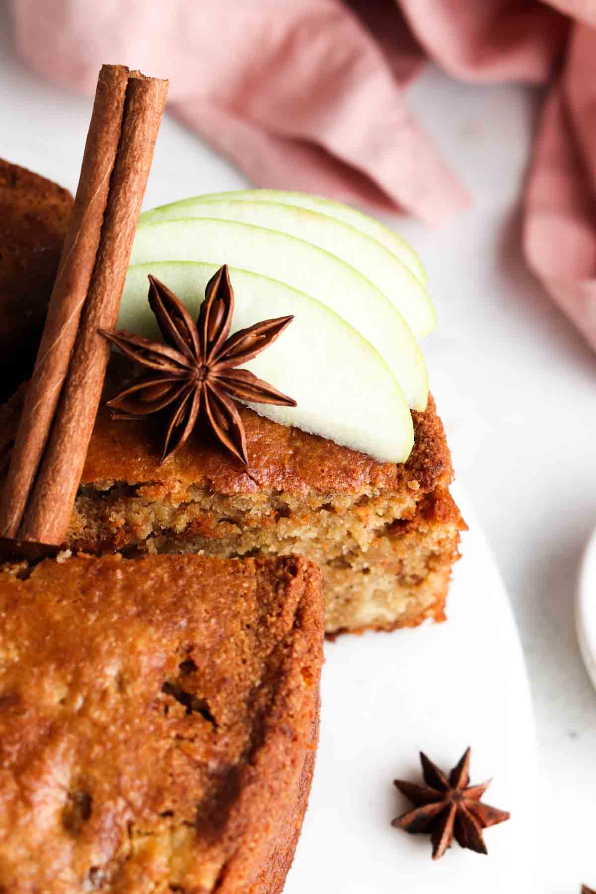 A slice of Gluten Free Apple Cake on a white and gray plate with green apples, a cinnamon stick and anise star on top.