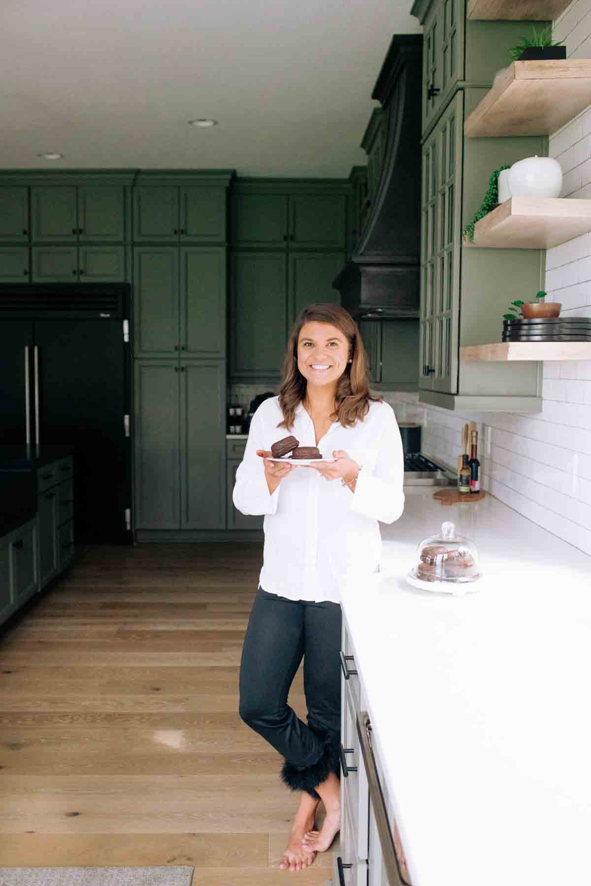 Picture of a woman in black pants and a white top standing next to the island holding a plate of donuts. 