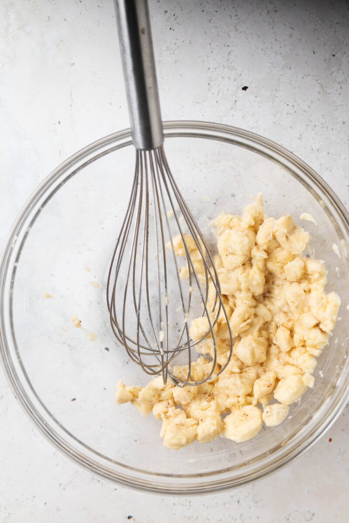 Banana mashed in a glass bowl with a whisk.