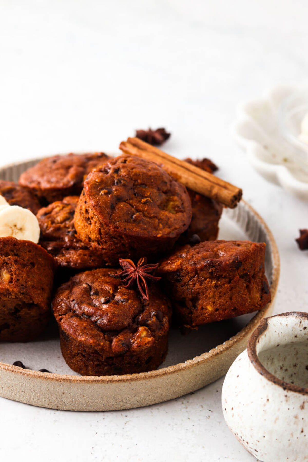 Gluten free pumpkin muffins stacked on a clay plate with whole star anise, cinnamon sticks, and fresh banana.