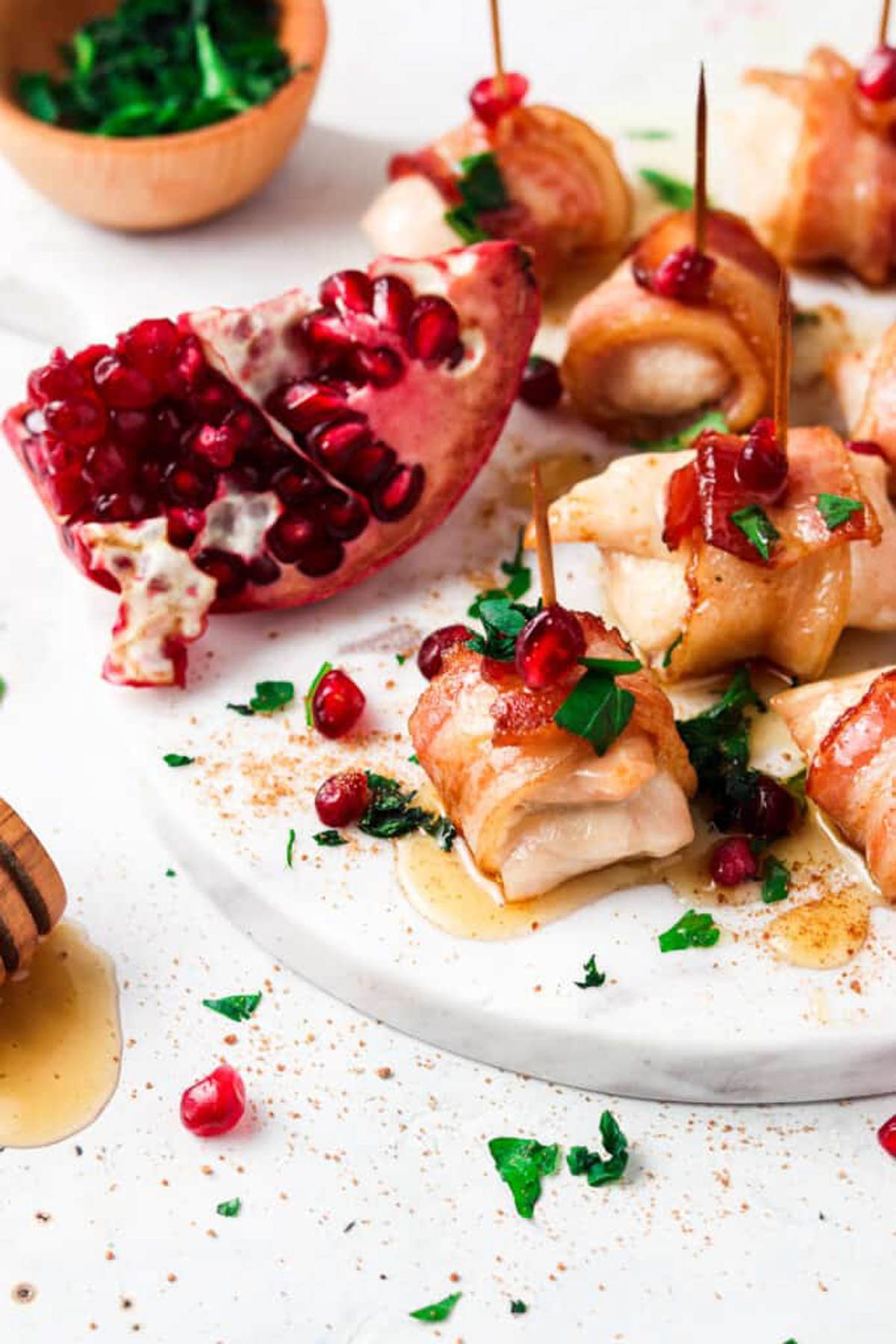 Bacon wrapped chicken bites drizzled with honey and garnished with fresh green herbs and pomegranate seeds on a marble platter. 
