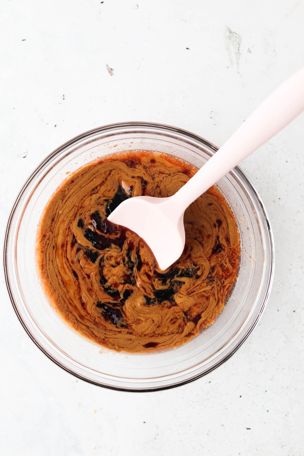 Peanut butter, lime juice, grape jelly, apple cider vinegar, sriracha, and ginger in a glass mixing bowl with a light pink spoon.