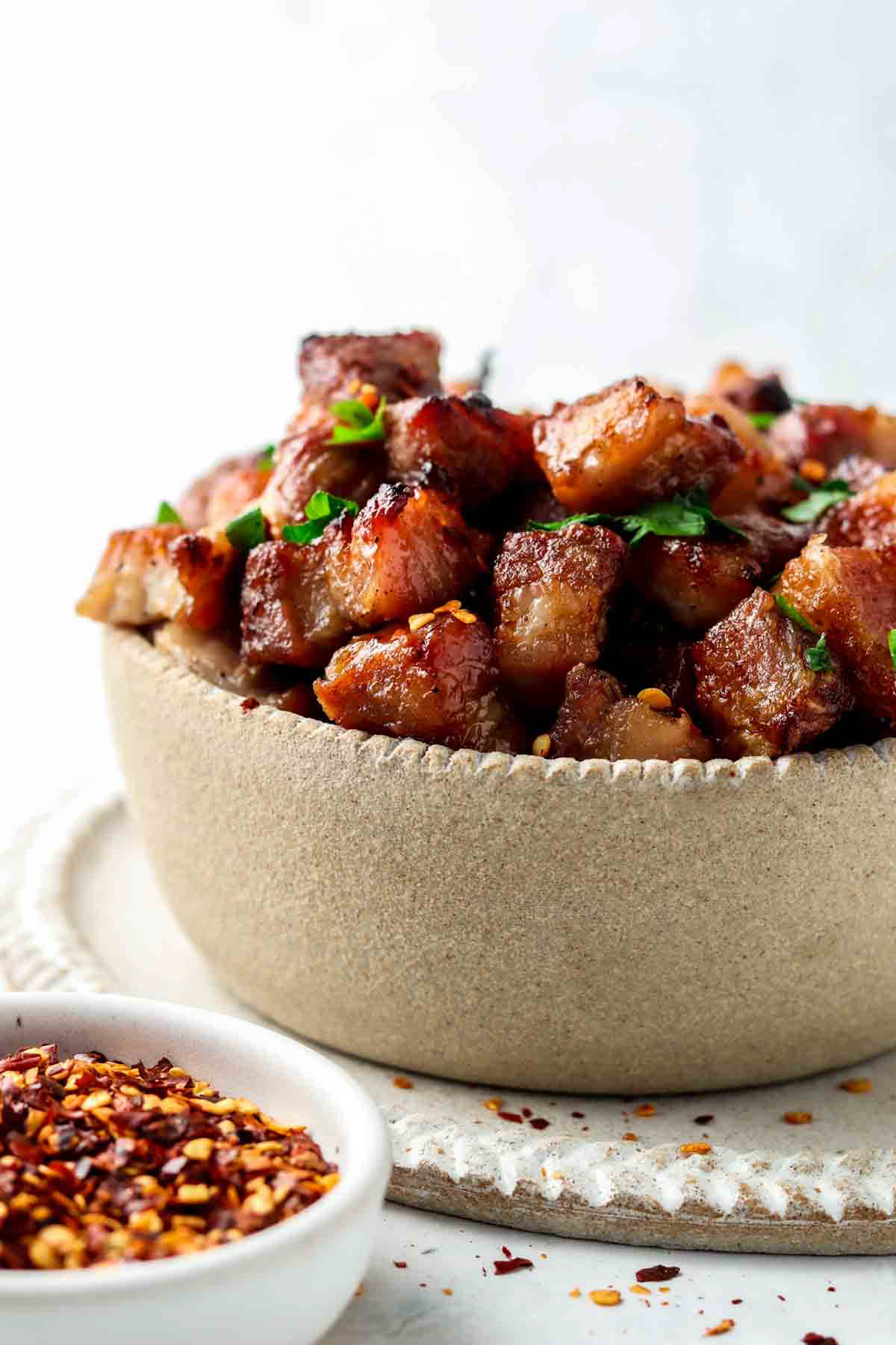 Air fryer pork belly in a clay bowl on a clay plate garnished with red pepper flakes and fresh green herbs.