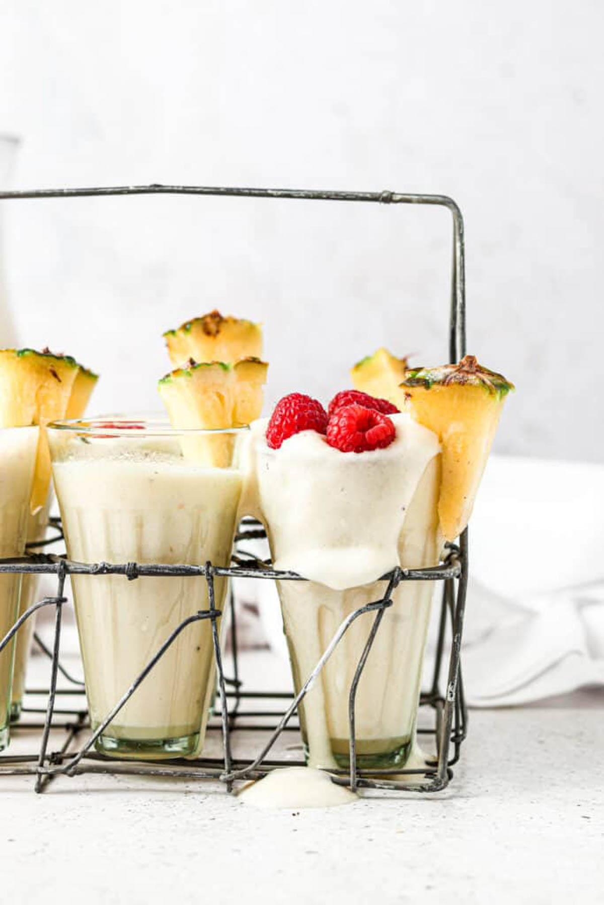 Six pine colada smoothies in class cups garnished with fresh pineapple wedges and raspberries.  