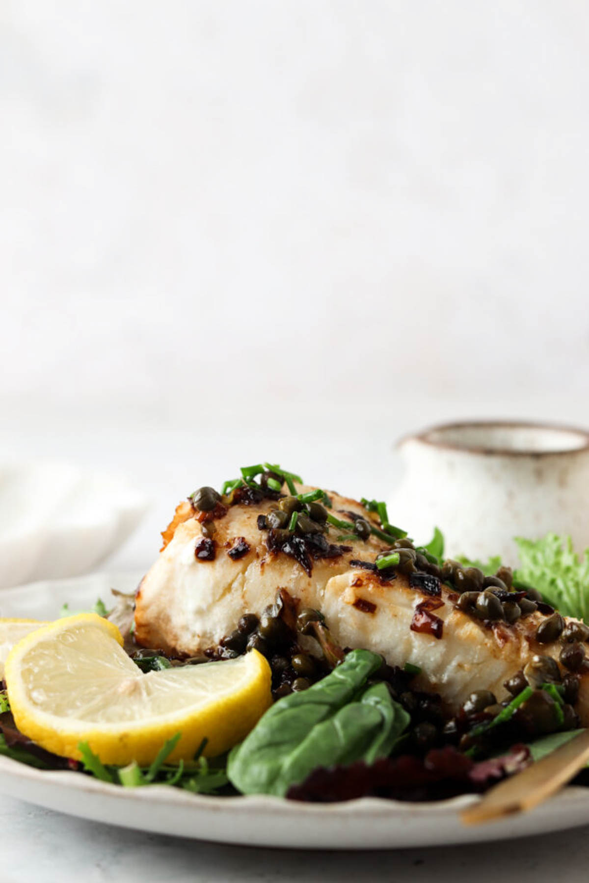 Pan seared chilean sea bass with lemon caper sauce on a bed of greens with a garnish of fresh lemon.