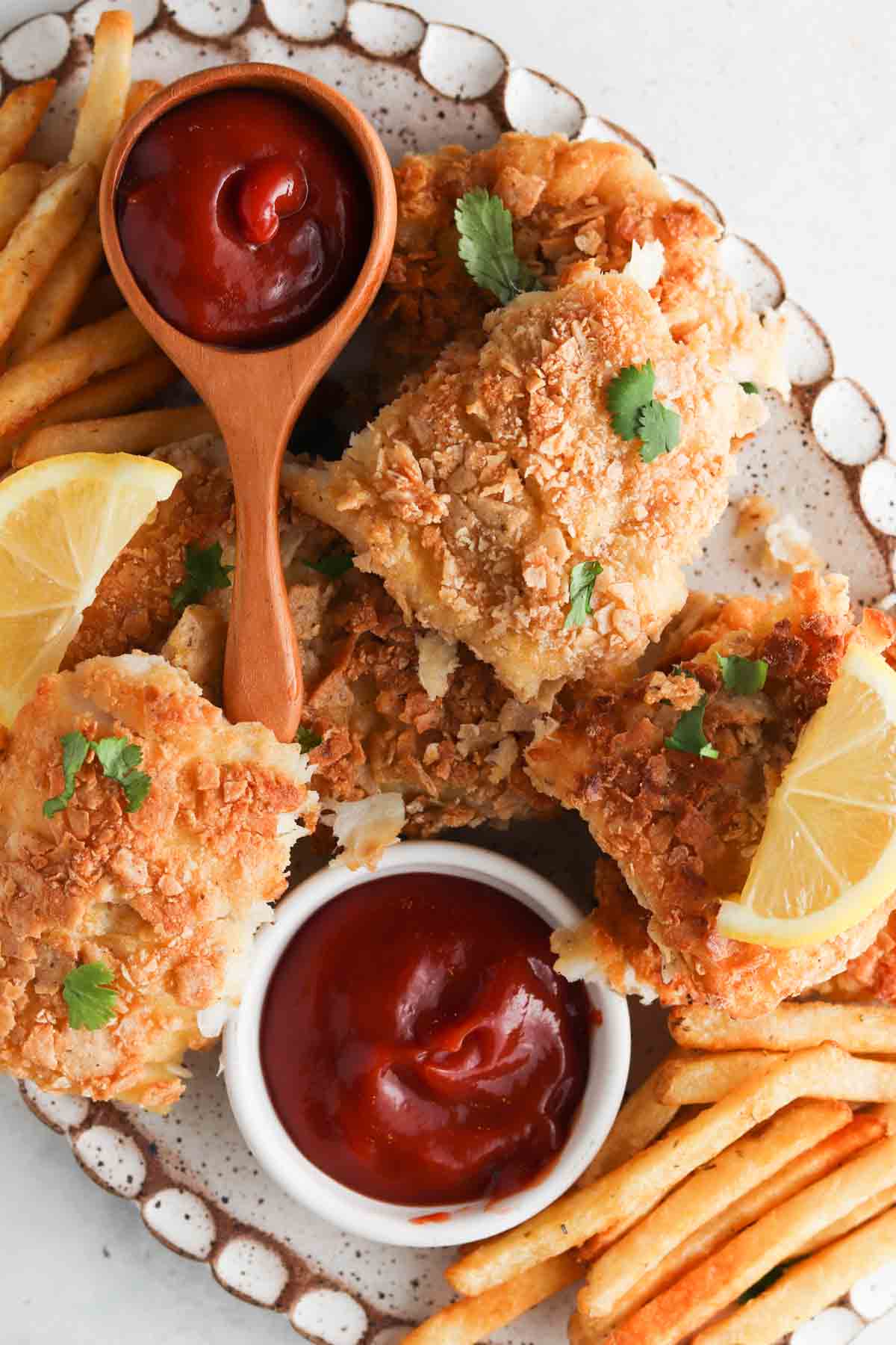 Gluten free fish and chips on a plate with ketchup, lemons, and fresh herbs.