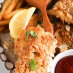 Close up photo of breaded fish on a plate with ketchup, french fries, lemon halves and fresh herbs.