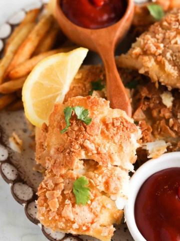 Close up photo of breaded fish on a plate with ketchup, french fries, lemon halves and fresh herbs.