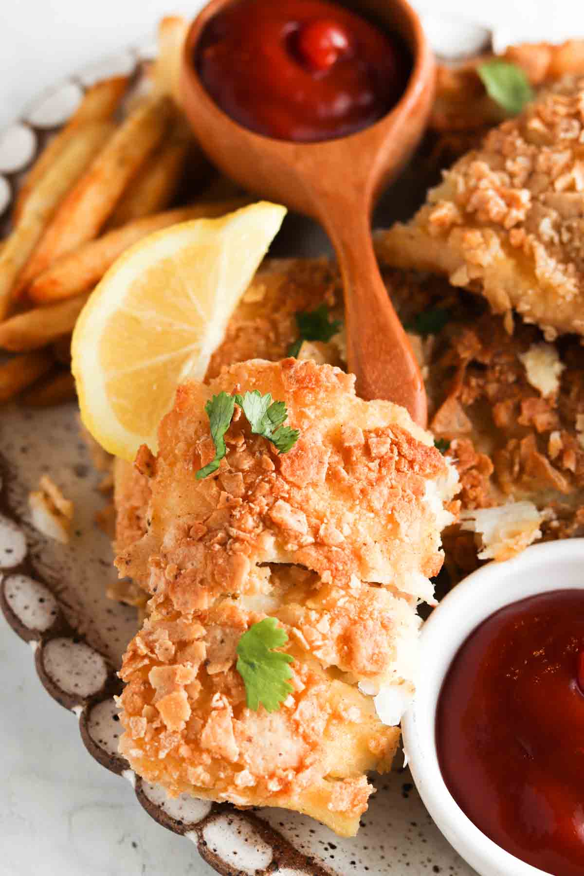 Close up photo of breaded fish on a plate with ketchup, french fries, lemon halves and fresh herbs.