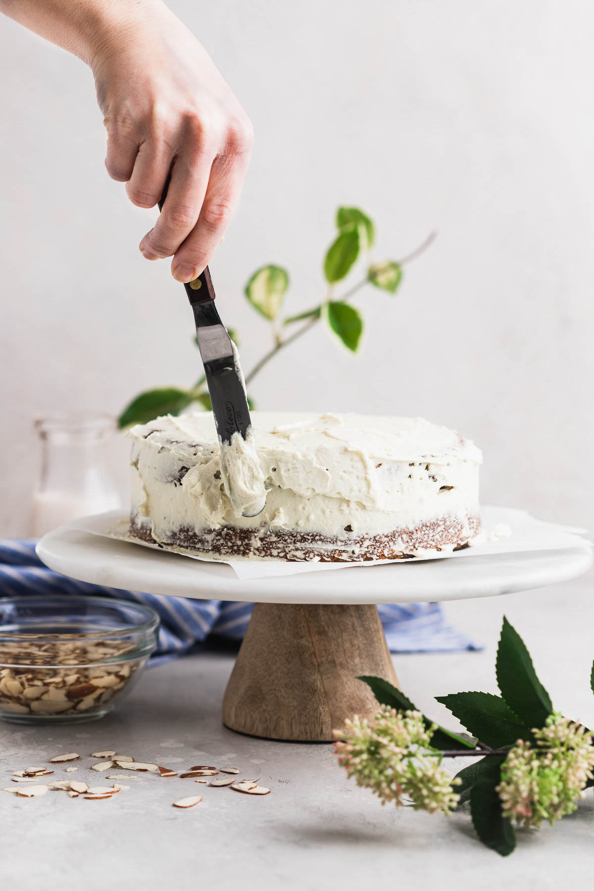 A person icing a carrot cake with a spatula on a pretty marble cake stand.