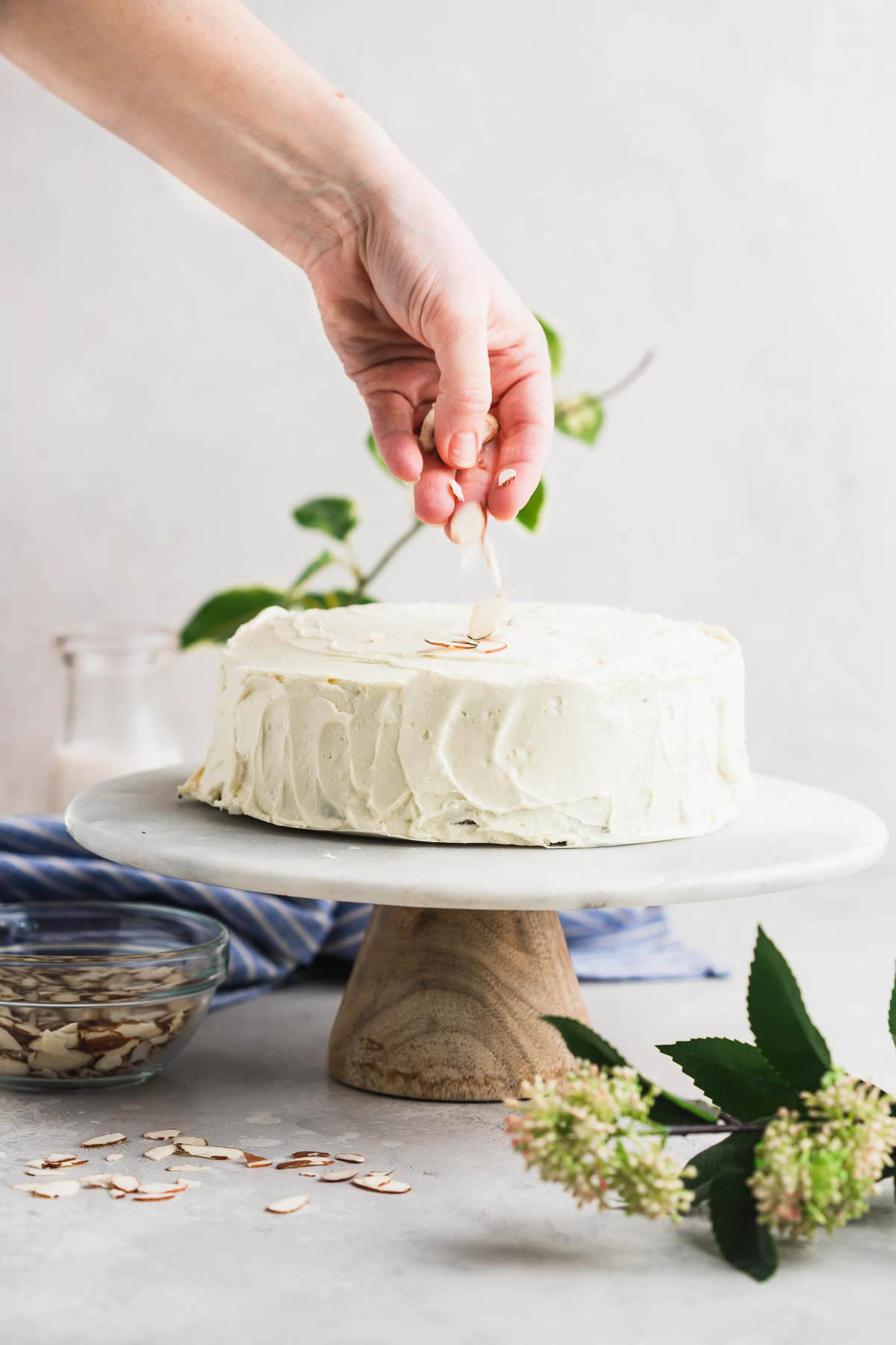 A person sprinkling fresh sliced almond slivers on top or an iced gluten free carrot cake. 