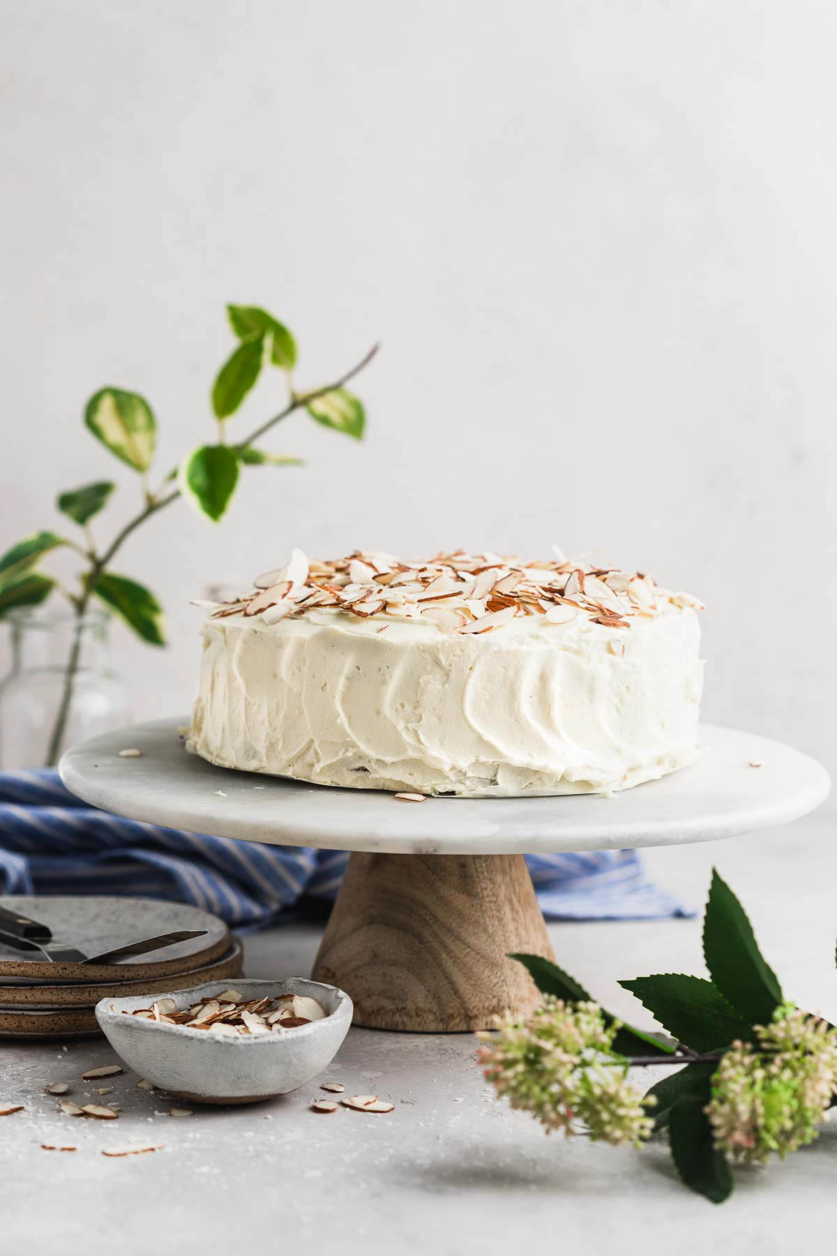 Carrot cake on a cake stand with almond slivers on top and green flowers on the counter. 