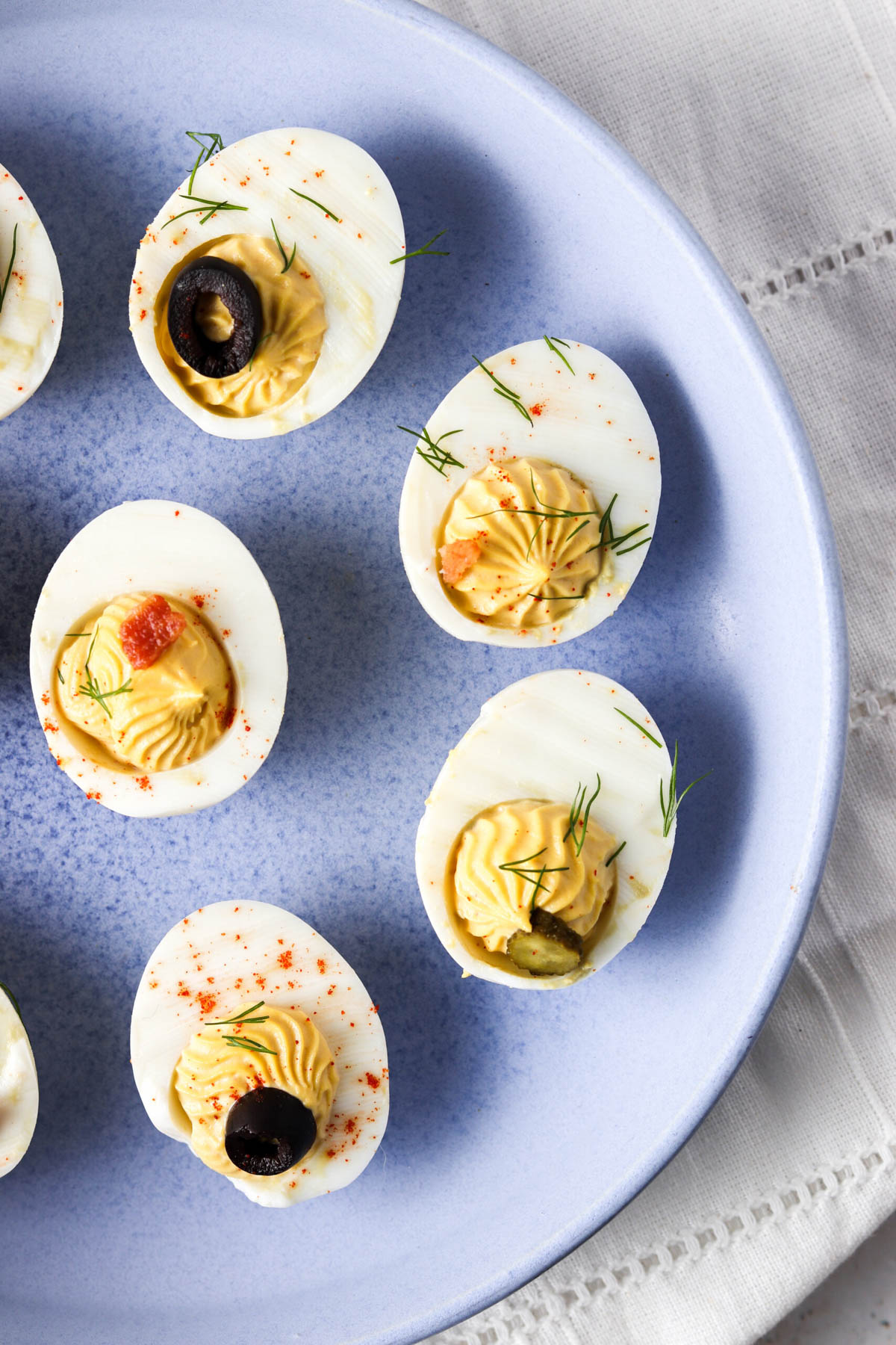 Deviled eggs on a plate with fresh herbs, olives, bacon and paprika on top.