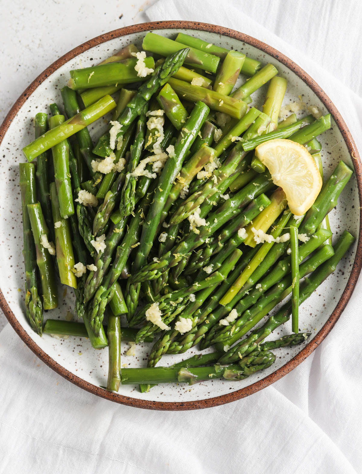 A platter of lemon garlic asparagus on a plate with fresh garlic and a lemon slice.