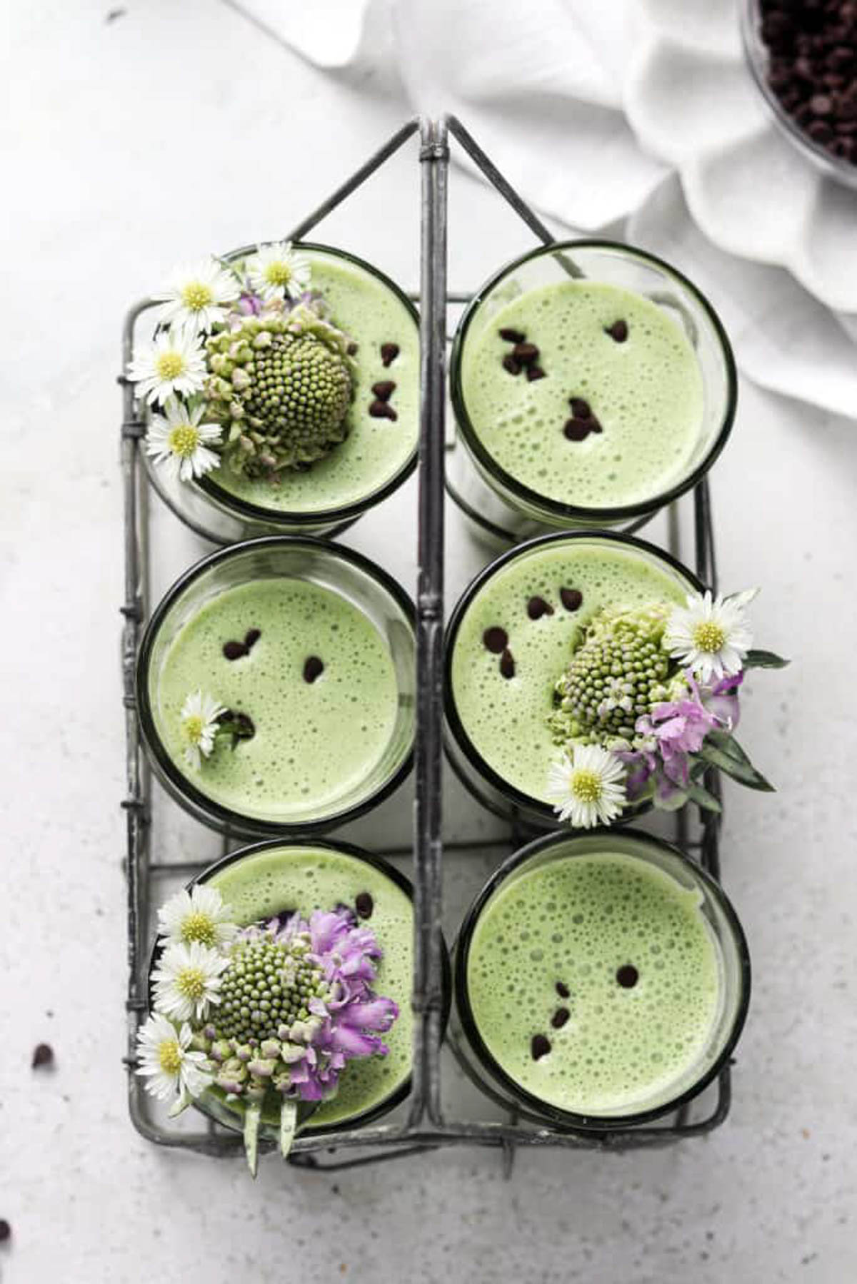 Six mint smoothies in a wire rack and garnished with fresh flowers and chocolate chips.