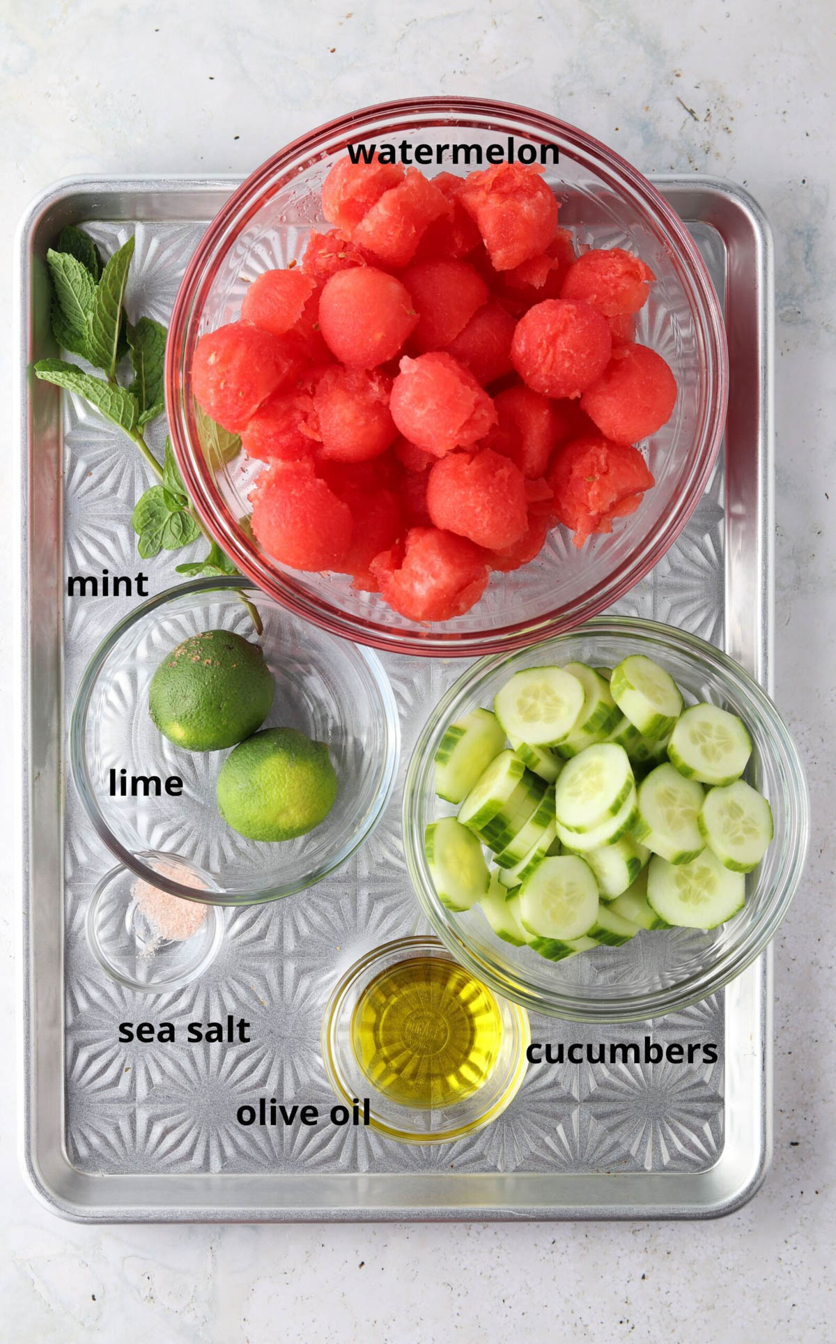 Cucumber watermelon salad ingredients in small glass bowls on a silver tray.