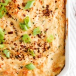 Crab dip in a white casserole dish with green onions and red pepper flakes on top.