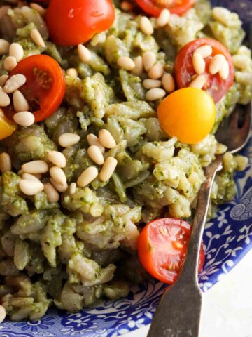 Close up photo of pesto orzo salad in a blue bowl with tomatoes and pine nuts on top.