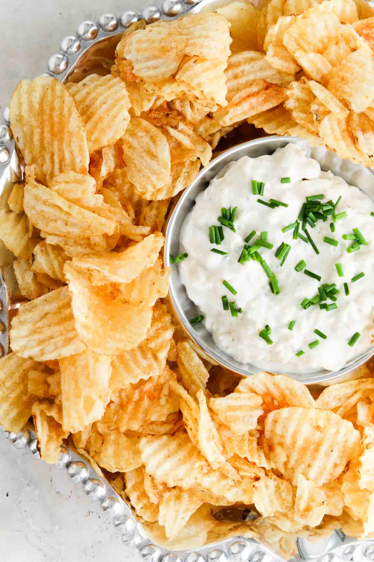 Pretty french onion dip in a silver bowl with potato chips and chives on top.