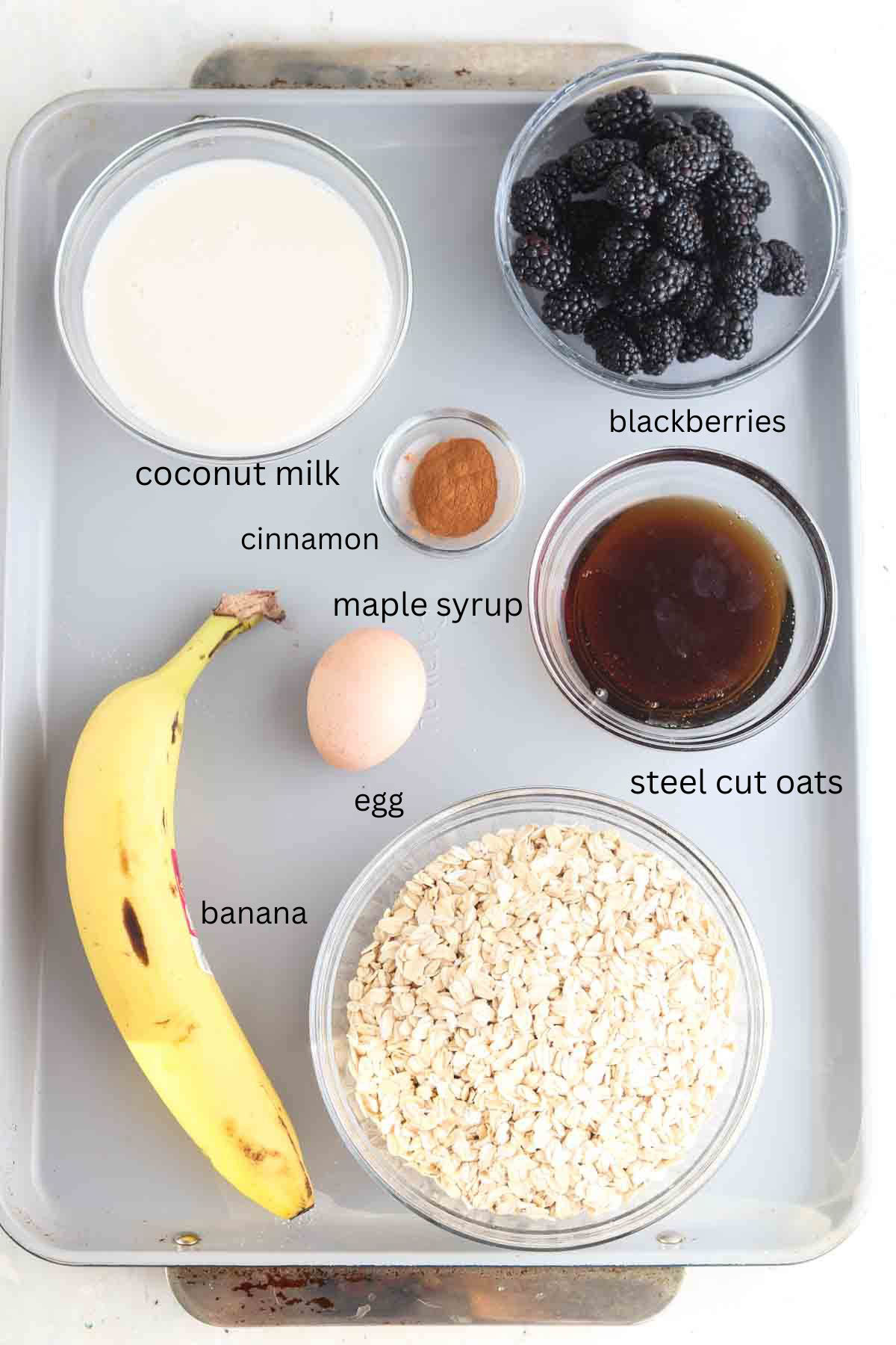 Oats, bananas, blackberries, maple syrup, cinnamon, milk and an egg laid out on the counter in glass clear bowls.