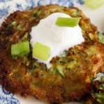 Close up photo of a zucchini fritter on a plate with sour cream and green onions on top.