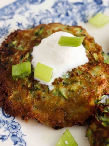 Close up photo of a zucchini fritter on a plate with sour cream and green onions on top.
