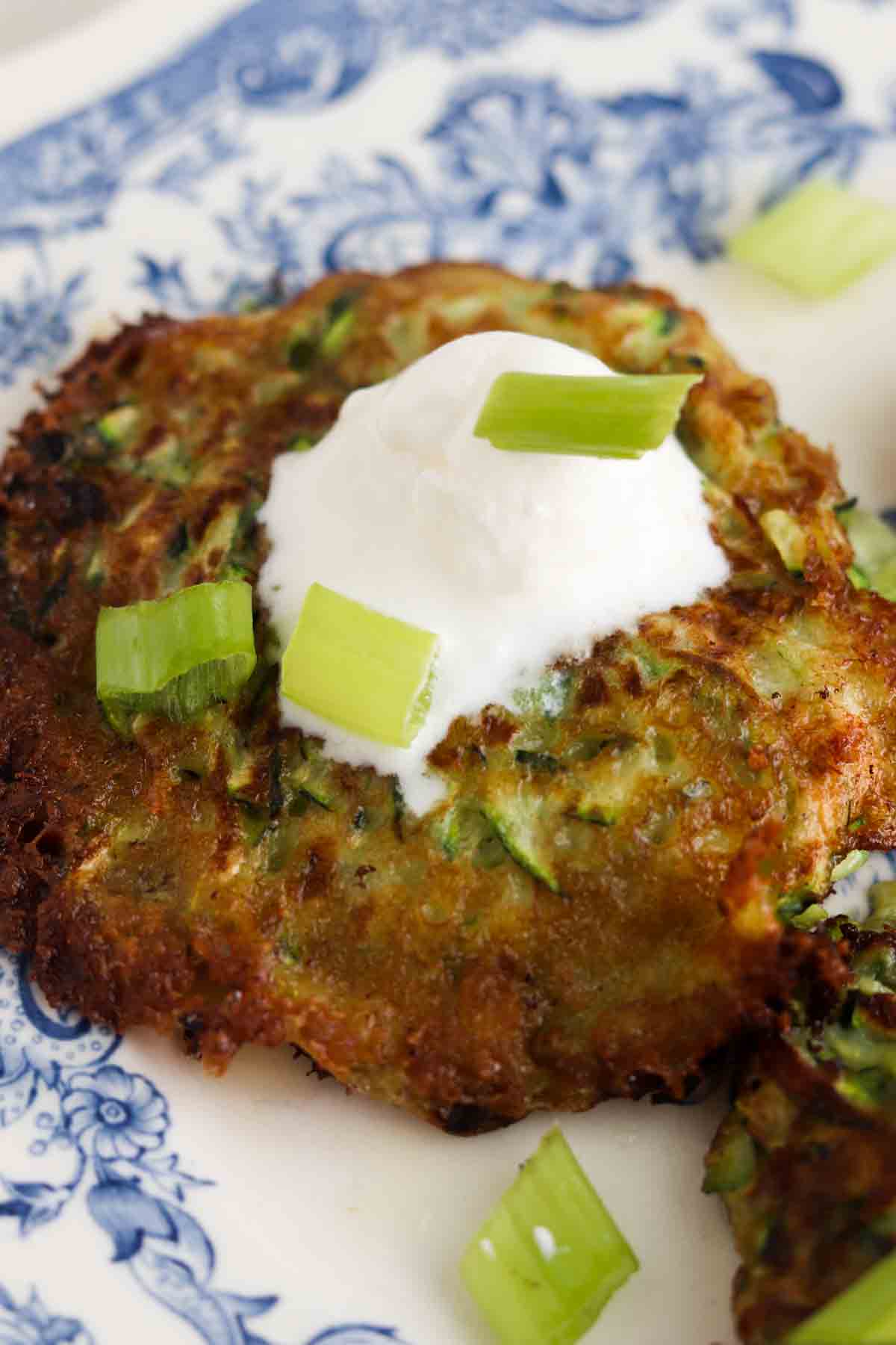 Close up photo of a zucchini fritter on a plate with sour cream and green onions on top.